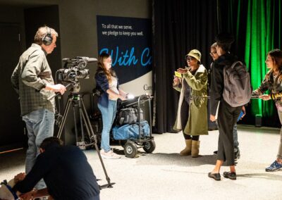 Film crew records talent in front of a sign while adjusting equipment and lighting.
