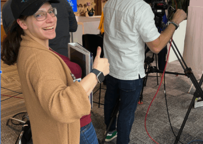 Crew member gives a thumbs up behind the scenes of a studio set with camera equipment and lighting.