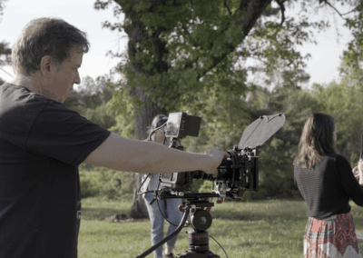 Cinematographer adjusts camera on a tripod during an outdoor shoot with a wooded backdrop.