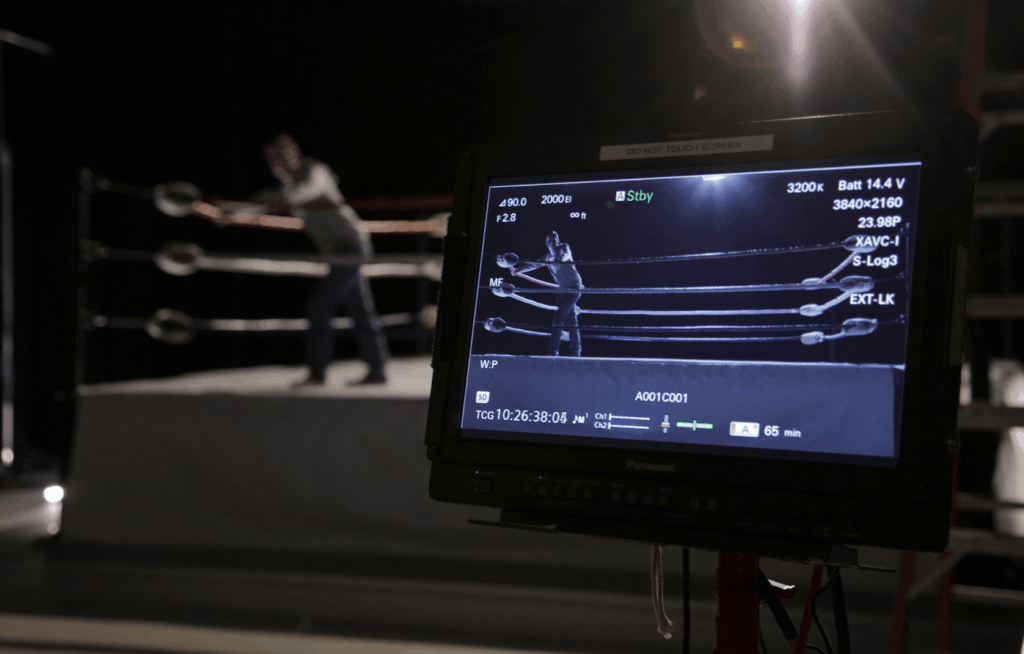 A studio camera monitor shows a scene inside a wrestling or boxing ring with a person standing between the ropes. The actual ring setup is visible in the background of the studio.