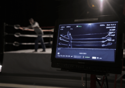 A studio camera monitor shows a scene inside a wrestling or boxing ring with a person standing between the ropes. The actual ring setup is visible in the background of the studio.